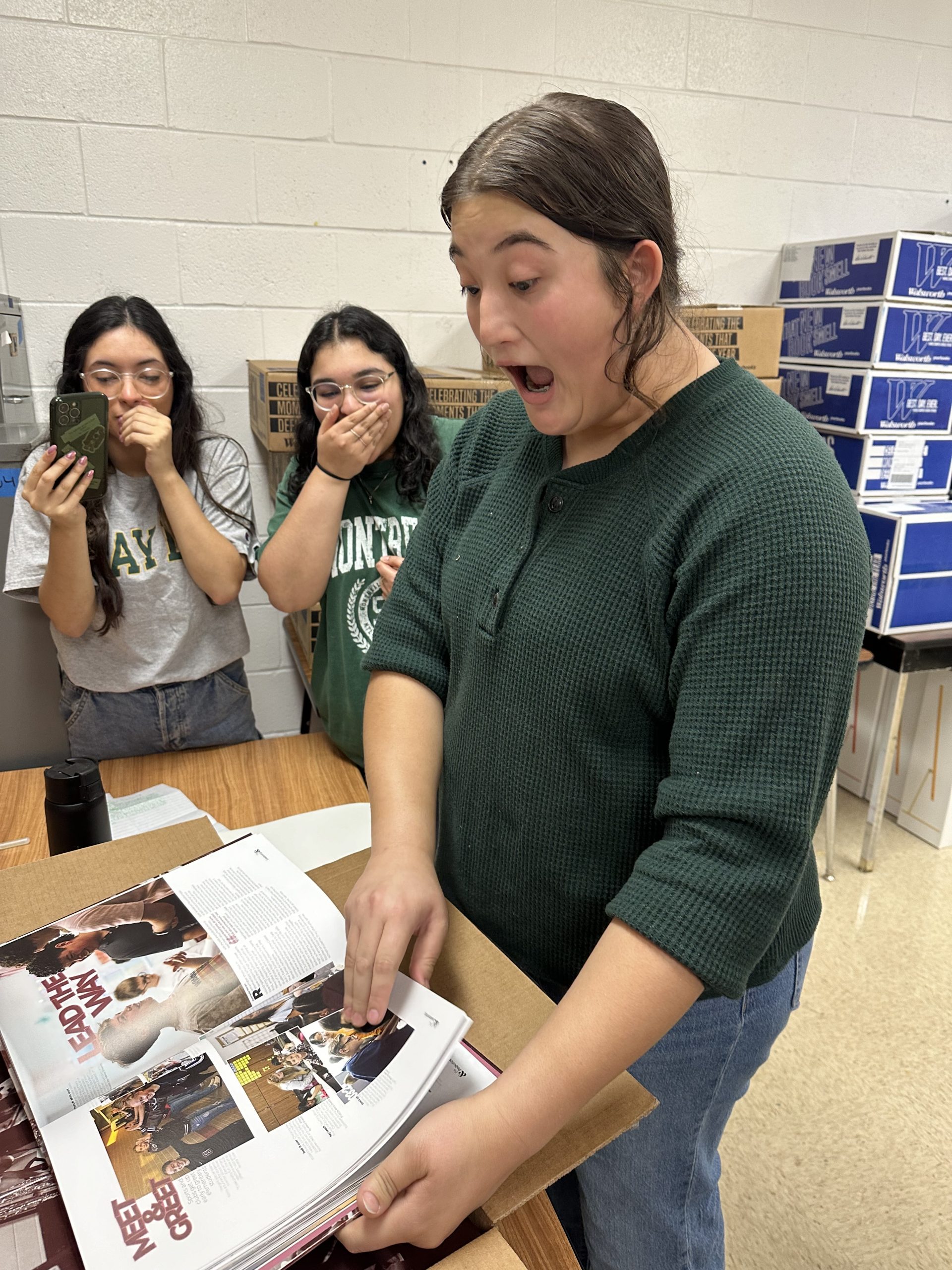 Three students standing around a yearbook pointing at a photo. Their expressions are in surprise. The photo in the yearbook that the students are pointing at is just a group photo of more students. Nothing to be alarmed by, trust me.
