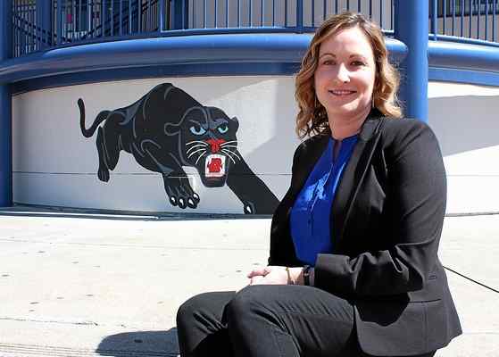 Brianna Kleinschmidt, a woman principal, sitting in front a painting of a panther on a wall in front of the school she is the principal of.