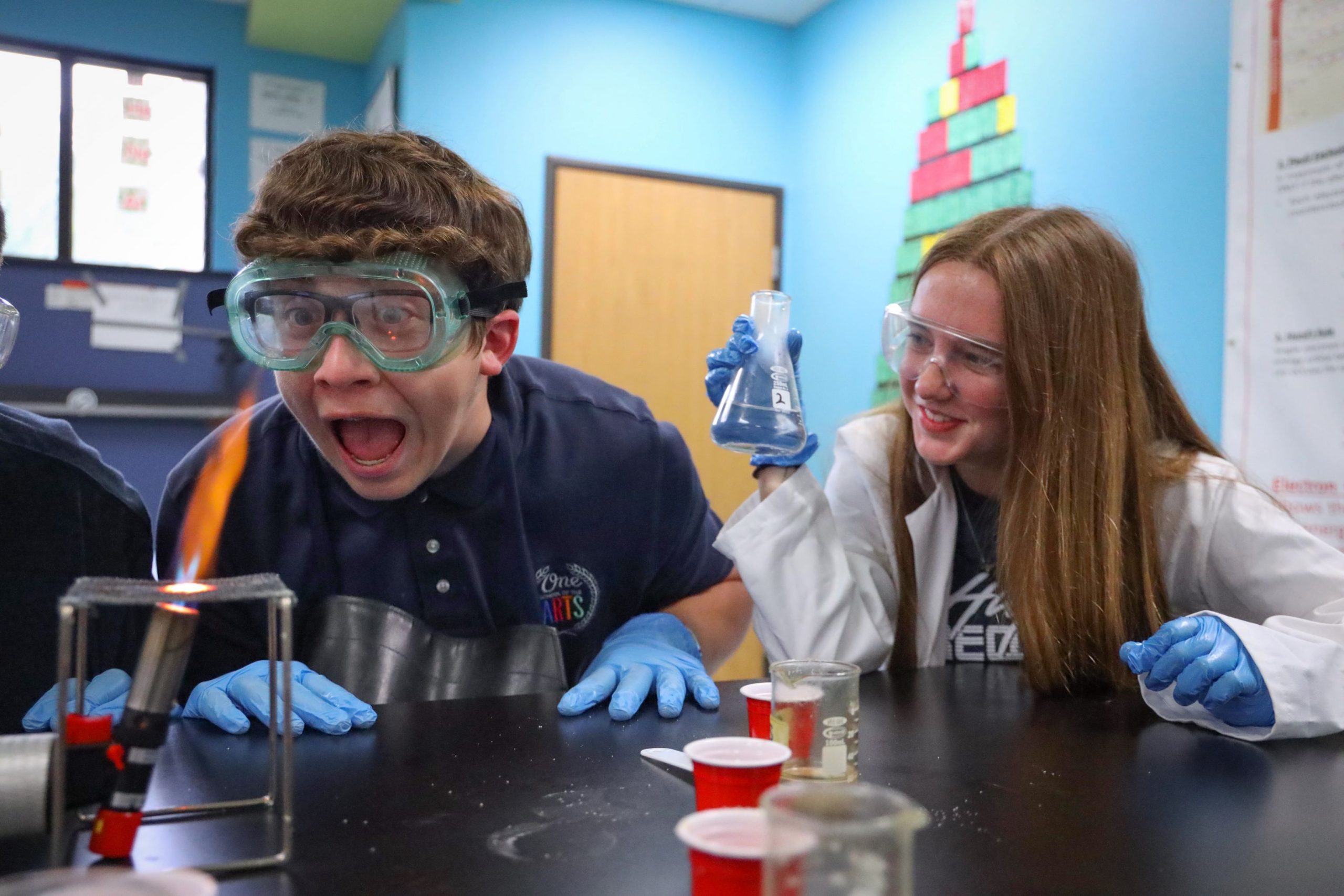a photo of two students in a science lab. One has a shocked expression looking at the science experiment and the other student is grinning at the other student's reaction.