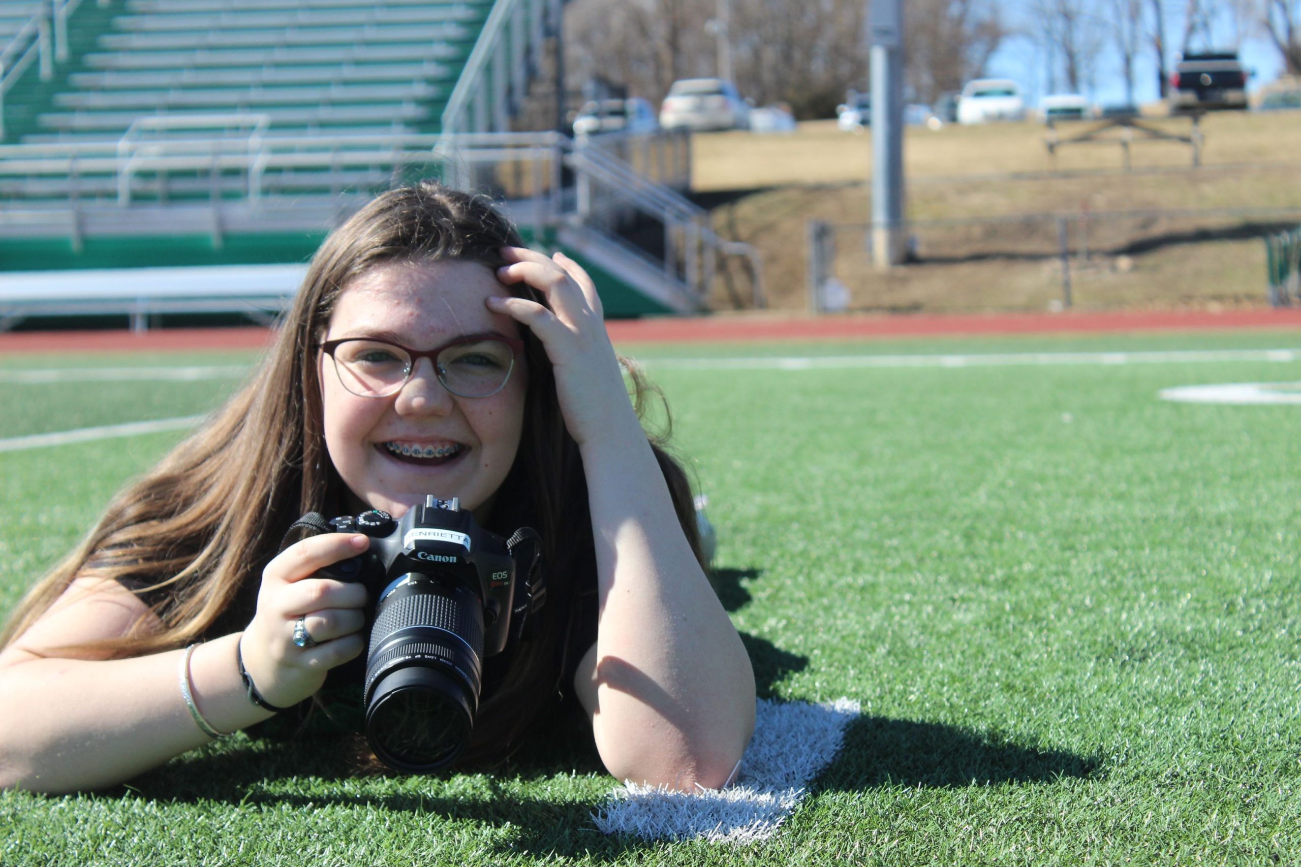 A girl laying on her stomach on a football field, holding a camera and smiling.