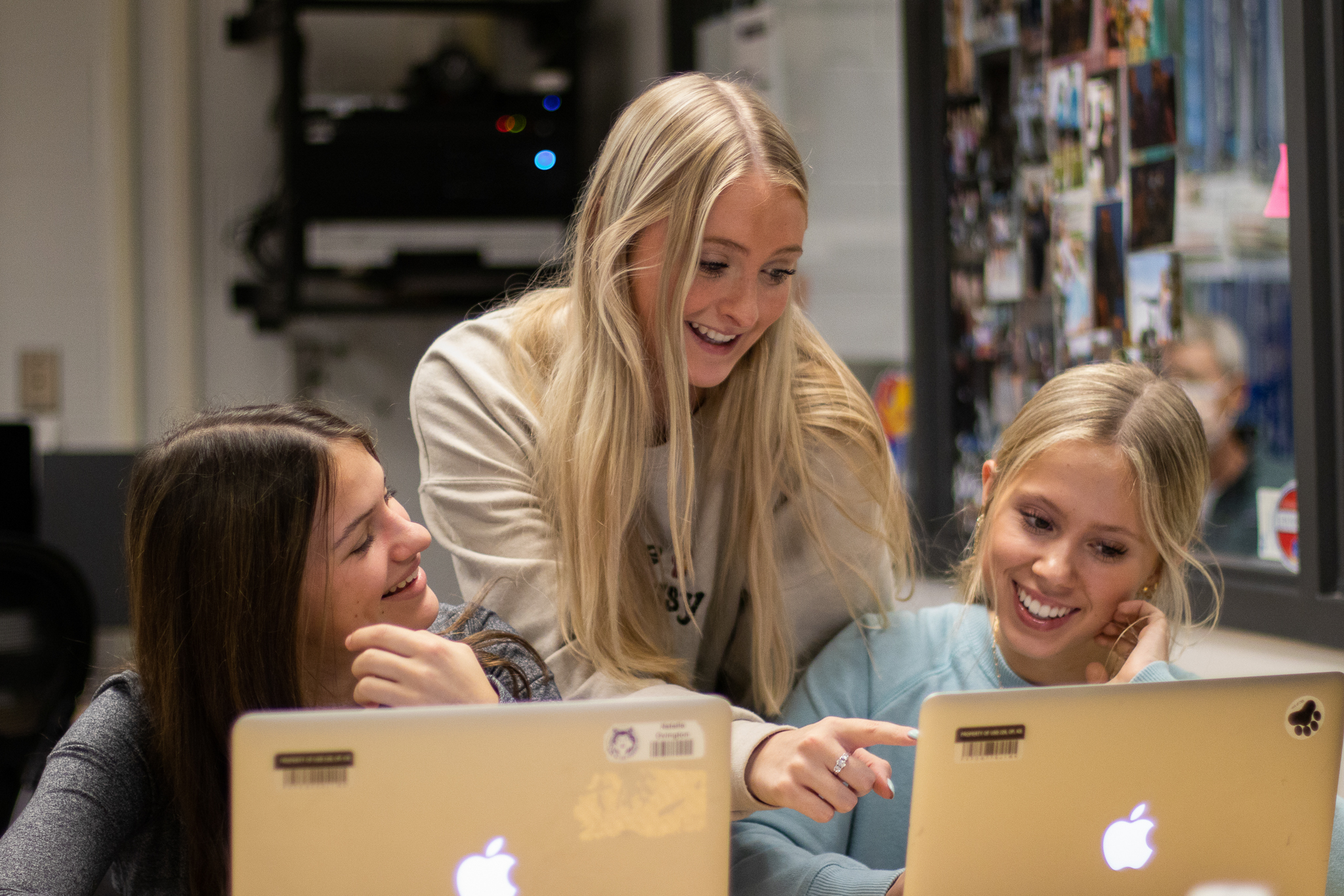 A photo of three girls looking at their computer screens. One girl is behind them and pointing at something on their screen. They are smiling and laughing.